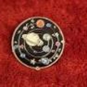 May include: A round, black enamel pin with a silver border. The pin features a cream-colored planet with rings, surrounded by smaller planets, stars, and floral accents. The design is set against a black background.
