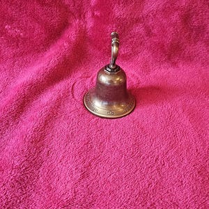 K&ouml;nnte beinhalten: Eine kleine, antike Bronzeglocke mit gebogenem Griff. Die Glocke hat einen ausgestellten Sockel mit gepr&auml;gtem Text und dekorativen Elementen. Die Glocke befindet sich vor einem strukturierten, rosa Hintergrund.