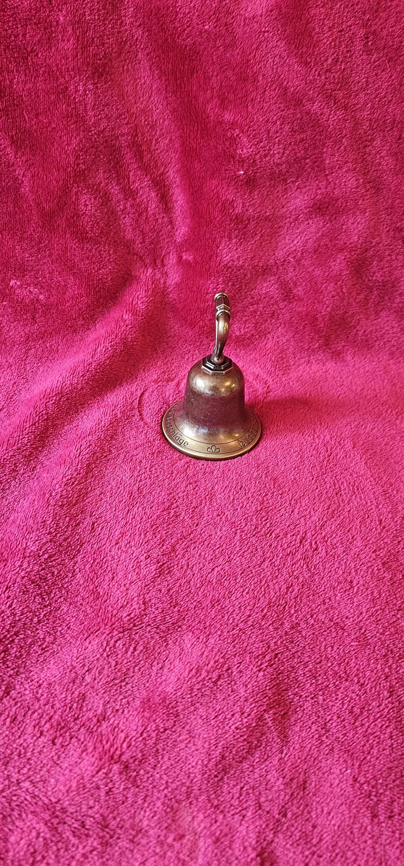 K&ouml;nnte beinhalten: Eine kleine, antike Bronzeglocke mit gebogenem Griff. Die Glocke hat einen ausgestellten Sockel mit gepr&auml;gtem Text und dekorativen Elementen. Die Glocke befindet sich vor einem strukturierten, rosa Hintergrund.