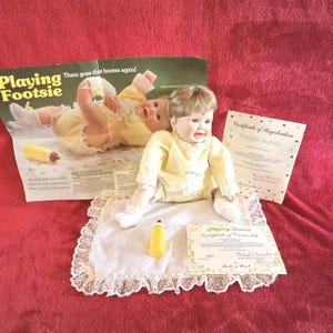 Könnte beinhalten: Porzellan-Babypuppe in einem gelben Outfit mit weißer Spitzenborte, sitzend auf einem weißen Tuch mit Spitzenkante. Eine gelbe Flasche und Papiere sind ebenfalls vorhanden. Die Puppe gehört zur "Playing Footsie"-Kollektion.