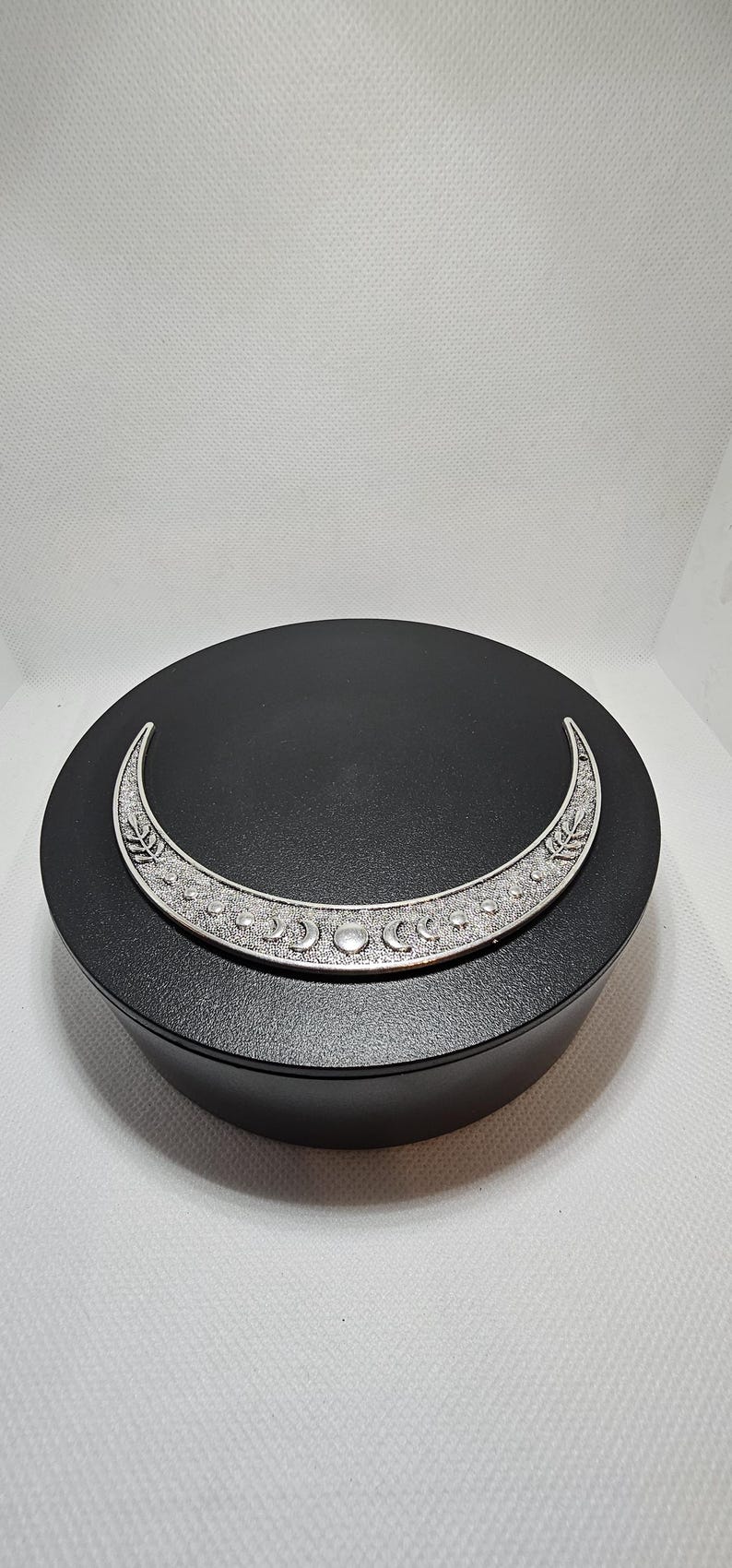 Puede incluir: Una caja redonda negra con un dise&ntilde;o de media luna plateada. La luna tiene una superficie texturizada y presenta una serie de lunas m&aacute;s peque&ntilde;as y un dise&ntilde;o de hojas.