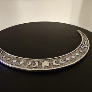 Puede incluir: Una luna creciente de plata con una superficie texturizada y un dise&ntilde;o de peque&ntilde;as lunas y hojas. La luna est&aacute; sobre una superficie negra.