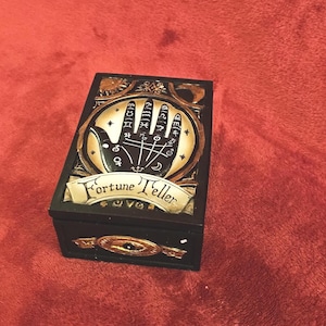 May include: A black rectangular box with a gold and cream-coloured palmistry design on the lid. The lid features a hand with lines and symbols, and the words "Fortune Teller" are written on a banner below the hand.