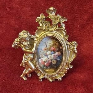May include: Ornate gold-colored picture frame with an oval-shaped floral painting. A small cherub figure is attached to the left side of the frame. The frame has decorative floral and scrollwork details. The background is a deep red color.