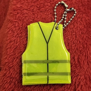 May include: A yellow safety vest keychain with black stripes and a black Y-shaped design. The keychain is attached to a silver ball chain. The vest is a bright, fluorescent yellow color.