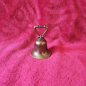 K&ouml;nnte beinhalten: Eine kleine, antike Messingglocke mit Griff, die auf einer rosa, strukturierten Oberfl&auml;che ruht. Die Glocke hat eine klassische Form und eine leicht angelaufene Oberfl&auml;che, was auf ihr Alter hindeutet. Die Basis der Glocke hat Text, der schwer zu lesen ist.