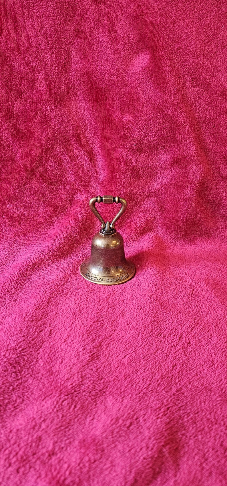 K&ouml;nnte beinhalten: Eine kleine, antike Messingglocke mit Griff, die auf einer rosa, strukturierten Oberfl&auml;che ruht. Die Glocke hat eine klassische Form und eine leicht angelaufene Oberfl&auml;che, was auf ihr Alter hindeutet. Die Basis der Glocke hat Text, der schwer zu lesen ist.