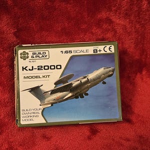 Könnte beinhalten: Modellbausatz-Box für das Flugzeug KJ-2000. Die Box zeigt ein Bild des Flugzeugs im Flug vor blauem Himmel. Auf der Box steht "Build & Play" und "Model Kit". Der Maßstab ist 1:65 und das empfohlene Alter ist 8+.