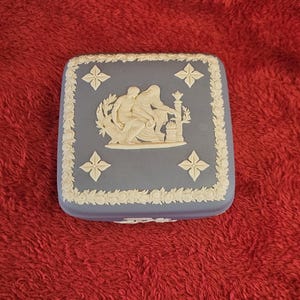 Könnte beinhalten: Quadratische blaue Schmuckschatulle mit weißem Reliefdesign klassischer Figuren und floralem Rand. Die Box hat ein dekoratives Kreuzmuster an jeder Ecke. Die Box befindet sich auf einer roten Oberfläche.
