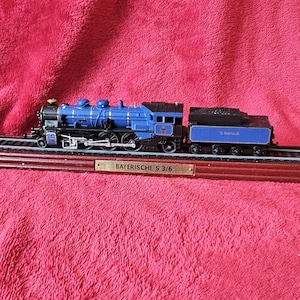 May include: A blue and black model train with a coal car, displayed on a wooden base with the text "BAYERISCHE S 3/6". The train has silver wheels and detailing. The background is a textured, red fabric.