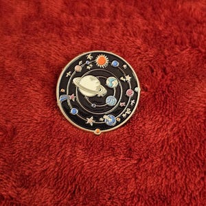 May include: A round, black enamel pin featuring a white planet with rings, surrounded by planets, stars, and a sun. The pin has a gold-colored border and is set against a red, textured background.