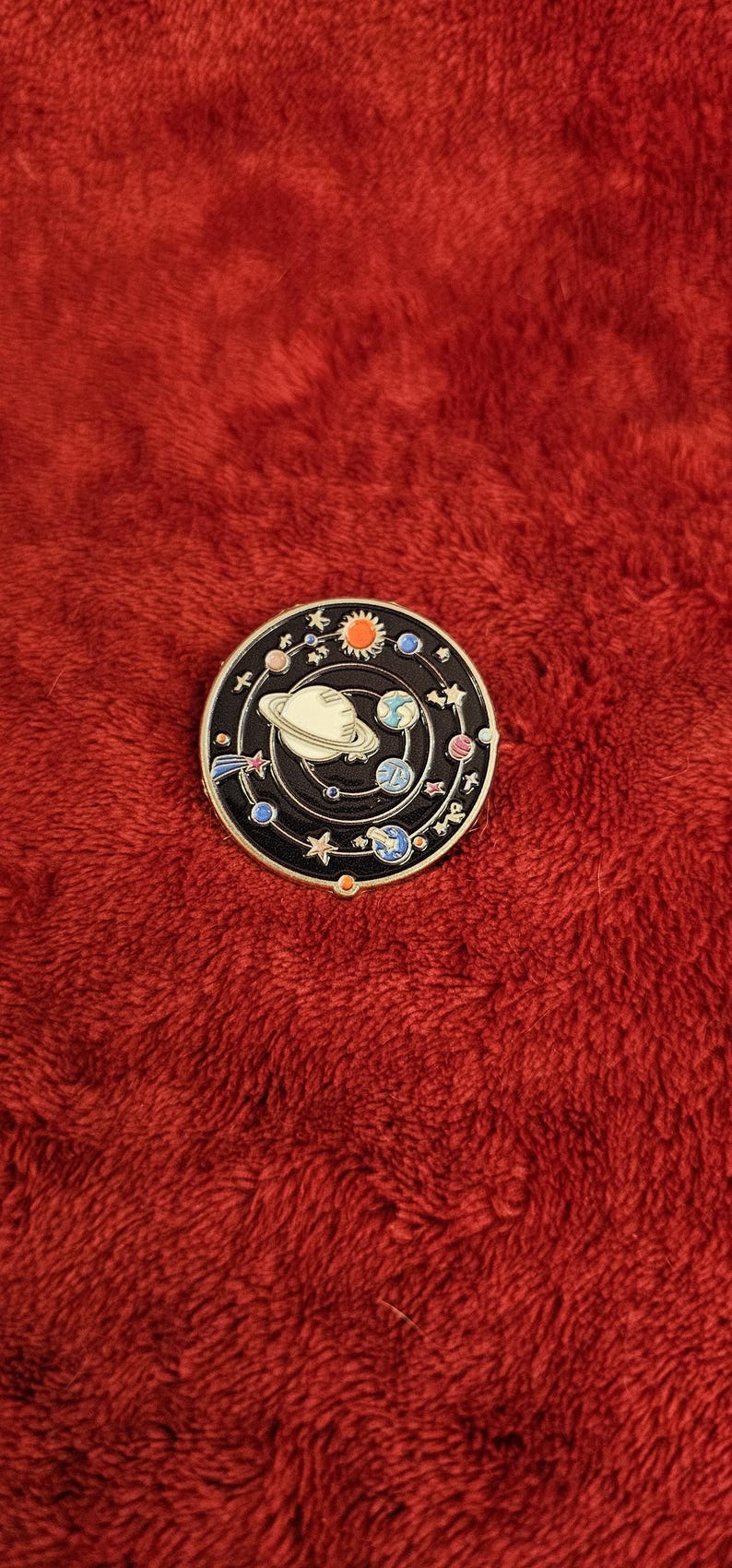May include: A round, black enamel pin featuring a white planet with rings, surrounded by planets, stars, and a sun. The pin has a gold-colored border and is set against a red, textured background.