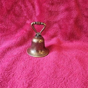 K&ouml;nnte beinhalten: Eine kleine Glocke im antiken Stil mit Bronze-Finish. Die Glocke hat einen abgerundeten K&ouml;rper und einen Griff. Der Sockel der Glocke hat Text. Die Glocke ist vor einem rosa Hintergrund platziert.