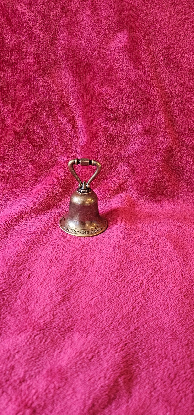 K&ouml;nnte beinhalten: Eine kleine Glocke im antiken Stil mit Bronze-Finish. Die Glocke hat einen abgerundeten K&ouml;rper und einen Griff. Der Sockel der Glocke hat Text. Die Glocke ist vor einem rosa Hintergrund platziert.