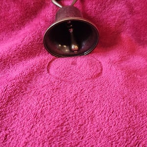 K&ouml;nnte beinhalten: Eine kleine Glocke im antiken Stil mit dunklem Metallfinish. Die Glocke hat einen Schlaufengriff und einen Kl&ouml;ppel im Inneren. Sie liegt auf einer strukturierten, fuchsiafarbenen Oberfl&auml;che.