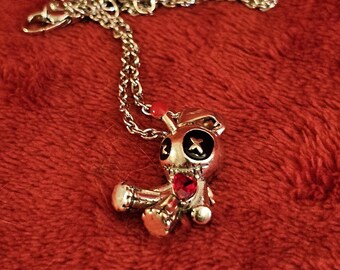 Può includere: Collana con pendente color argento con un design a bambola voodoo. La bambola ha occhi neri con una stella, un cuore rosso e una gemma rossa sulla testa. Su uno sfondo rosso strutturato.