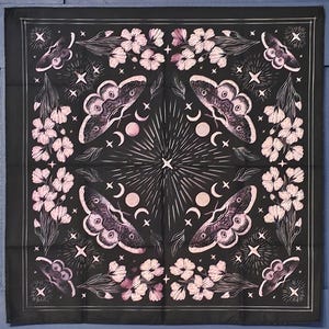 Puede incluir: Bandana negra con un diseño blanco con polillas, flores y lunas. El diseño es simétrico y tiene una estética vintage.