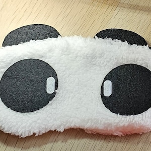Puede incluir: Una máscara para dormir con forma de panda, blanca y negra. La máscara presenta un exterior blanco y esponjoso con grandes círculos negros para los ojos y pequeñas orejas negras. Los círculos de los ojos tienen pequeños reflejos blancos. Se adjunta una banda elástica negra.