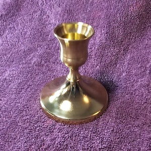 Könnte beinhalten: Ein goldfarbener Kerzenhalter aus Metall mit runder Basis und einem becherförmigen Oberteil. Der Halter ist aus poliertem Messing gefertigt und hat eine glatte, glänzende Oberfläche.