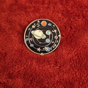May include: A round, black enamel pin featuring a solar system design. The pin includes planets, stars, and a sun, with a white Saturn in the center. The pin is set against a red, textured background.