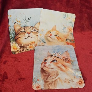 Puede incluir: Tres posavasos cuadrados con ilustraciones de acuarela de gatos sonrientes. Un posavasos presenta un gato atigrado rodeado de margaritas. Otro muestra un gato naranja con los ojos cerrados. El tercero tiene un gato con una corona de flores.