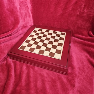 Puede incluir: Un juego de ajedrez de madera con un marco burdeos y una superficie de juego a cuadros. Los cuadrados alternan entre madera clara y marrón oscuro. El conjunto está cerrado, mostrando las capas apiladas del tablero.