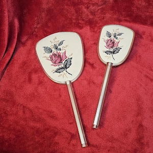 Könnte beinhalten: Zwei Vintage-Handspiegel mit silberfarbenen Griffen und ovalen Gesichtern. Jeder Spiegel hat einen cremefarbenen Hintergrund mit einem Design aus rosa Rosen und grünen Blättern. Die Spiegel sind vor einem roten Samthintergrund platziert.