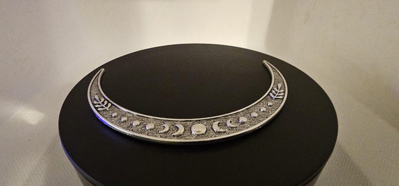 Puede incluir: Una luna creciente de plata con una superficie texturizada y un dise&ntilde;o de peque&ntilde;as lunas y hojas. La luna est&aacute; sobre una superficie negra.