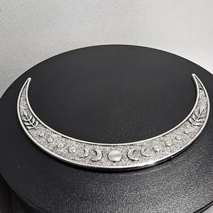 Puede incluir: Un colgante de plata en forma de media luna con un dise&ntilde;o detallado de lunas m&aacute;s peque&ntilde;as y hojas. El colgante est&aacute; sobre un fondo negro.