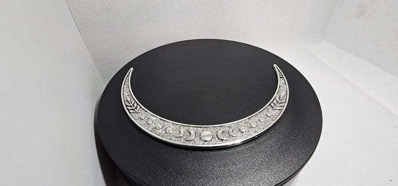 Puede incluir: Un colgante de plata en forma de media luna con un dise&ntilde;o detallado de lunas m&aacute;s peque&ntilde;as y hojas. El colgante est&aacute; sobre un fondo negro.
