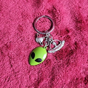 Könnte beinhalten: Ein silberfarbener Metallschlüsselanhänger mit einem runden Ring und drei Anhängern: einem kleinen silbernen Planeten mit Ringen, einem hellgrünen Alien-Kopf mit schwarzen Augen und einer silbernen fliegenden Untertasse mit der Gravur I WANT TO BELIEVE.