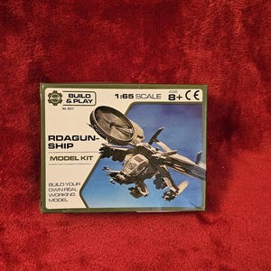 Puede incluir: Caja de modelo del RDAGUN-SHIP a escala 1:65. La caja presenta una ilustración detallada de la nave espacial contra un cielo azul. El texto dice «BUILD & PLAY» y «MODEL KIT». Recomendado para mayores de 8 años.
