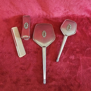 Könnte beinhalten: Ein silberfarbenes Vintage-Frisier-Set mit Kamm, Bürste und zwei Handspiegeln. Jedes Stück hat ein ovales Design. Die Artikel sind auf einer roten Oberfläche angeordnet.