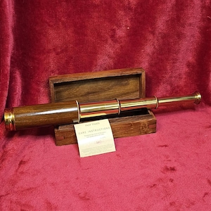 Könnte beinhalten: Ein Teleskop aus Messing und Holz im Vintage-Stil, das in einer offenen Holzkiste liegt. Das Teleskop hat eine polierte Messingoberfläche und einen Holzschaft. Eine kleine Karte mit Text ist sichtbar. Die Kiste und das Teleskop liegen auf rotem Untergrund.