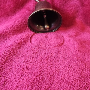 K&ouml;nnte beinhalten: Eine kleine Glocke im antiken Stil mit dunkler Bronzefinish. Die Glocke hat eine abgerundete Form mit einem Kl&ouml;ppel im Inneren und einem Griff oben. Sie liegt auf einer strukturierten, fuchsiafarbenen Oberfl&auml;che.