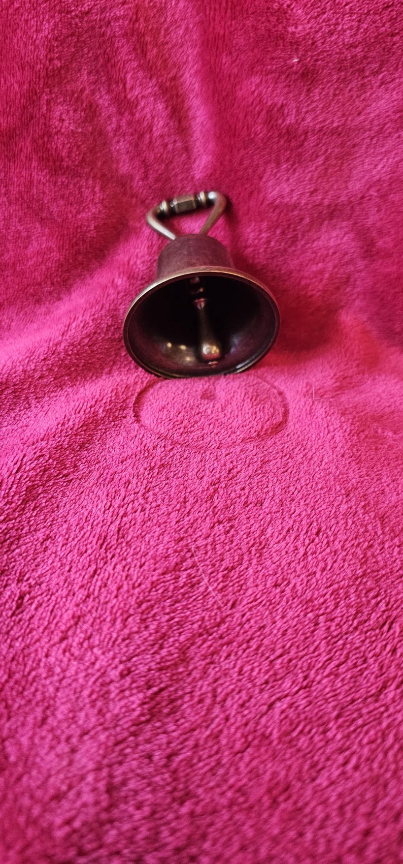 K&ouml;nnte beinhalten: Eine kleine Glocke im antiken Stil mit dunkler Bronzefinish. Die Glocke hat eine abgerundete Form mit einem Kl&ouml;ppel im Inneren und einem Griff oben. Sie liegt auf einer strukturierten, fuchsiafarbenen Oberfl&auml;che.