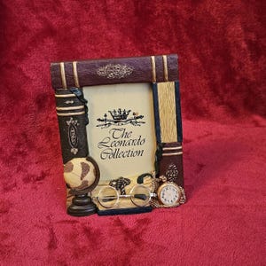 Peut inclure: Un cadre photo décoratif en forme de livre. Le cadre est marron et doré, avec un petit globe, des lunettes rondes et une montre de poche. Le texte "The Leonardo Collection" est visible sur le cadre.