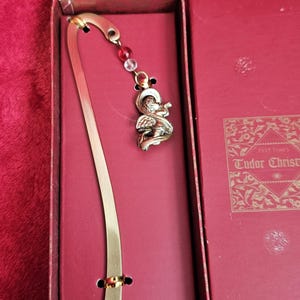 Könnte beinhalten: Ein goldfarbener Metall-Lesezeichen mit einem Engelsanhänger und roten und klaren Perlen, präsentiert in einer bordeauxfarbenen Schachtel. Das Lesezeichen hat ein geschwungenes Design mit einem Ringdetail. Die Schachtel trägt den Text "Tudor Christmas".