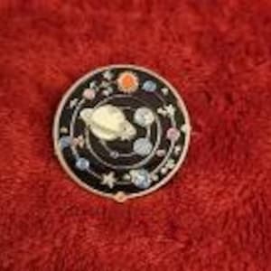 May include: A round, black enamel pin with a gold-colored planet and orbiting planets, stars, and other celestial bodies. The pin has a gold-colored border and is set against a red background.