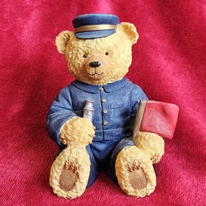 Könnte beinhalten: Dekorative Teddybär-Figur in blauer Uniform und Mütze, die einen silbernen Gegenstand und eine rote Flagge hält. Der Bär hat hellbraunes Fell und sitzt auf rotem Grund. Eine Sammlerfigur.