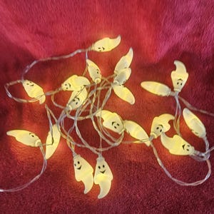 May include: A string of Halloween-themed lights featuring small, smiling ghost-shaped bulbs. The lights are a warm, soft yellow and are strung on a clear wire. The background is a deep red, textured fabric.