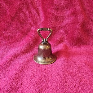 K&ouml;nnte beinhalten: Eine kleine Glocke im antiken Stil mit Bronze-Finish. Die Glocke hat einen Griff und einen ausgestellten Sockel, mit dem Text "zucker-beschl&auml;ge" am unteren Rand. Die Glocke befindet sich vor einem rosa Hintergrund.