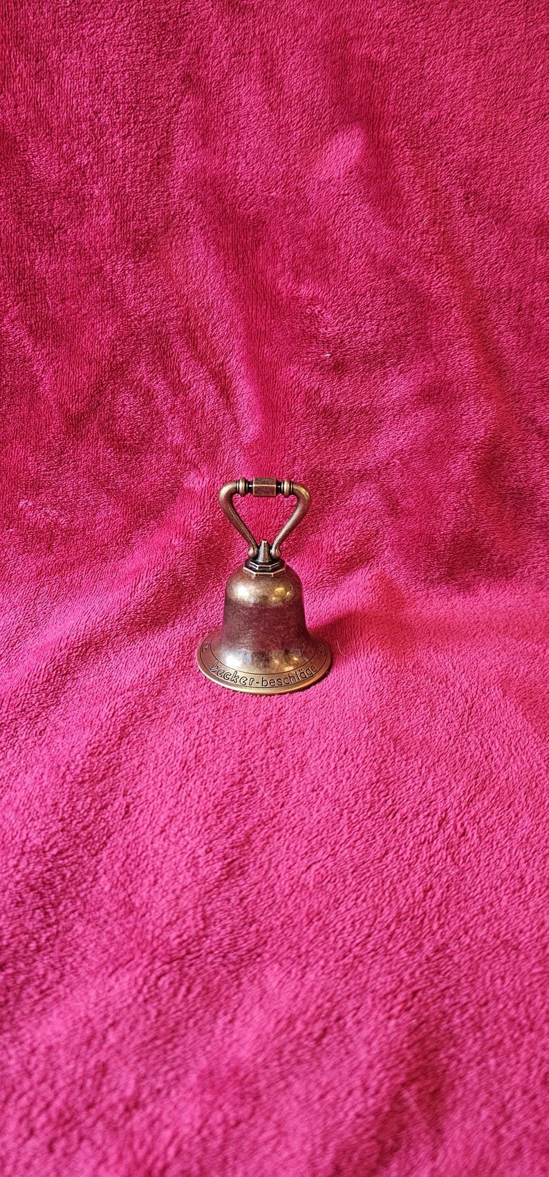 K&ouml;nnte beinhalten: Eine kleine Glocke im antiken Stil mit Bronze-Finish. Die Glocke hat einen Griff und einen ausgestellten Sockel, mit dem Text "zucker-beschl&auml;ge" am unteren Rand. Die Glocke befindet sich vor einem rosa Hintergrund.