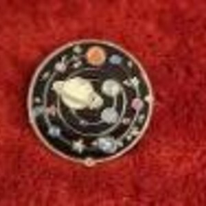 May include: A round, black enamel pin with a silver border. The pin features a cream-colored planet with rings, surrounded by smaller planets, stars, and floral accents. The design is set against a black background.