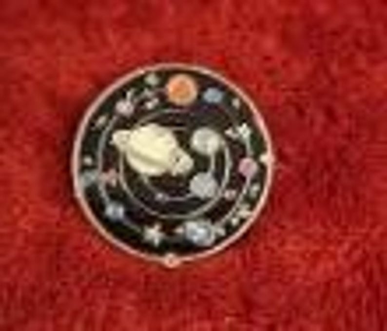 May include: A round, black enamel pin with a silver border. The pin features a cream-colored planet with rings, surrounded by smaller planets, stars, and floral accents. The design is set against a black background.