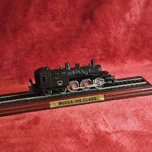 Könnte beinhalten: Ein schwarzes Modell der MUCCA 500 CLASS-Lokomotive steht auf einem Holzsockel mit Gleisen. Der Zug hat rot detaillierte Räder und ist vor einem roten Hintergrund platziert. Das Modell ist eine detaillierte Nachbildung.