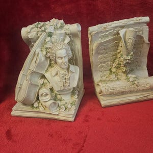 Könnte beinhalten: Zwei cremefarbene Buchstützen mit musikalischem Thema. Eine zeigt eine Büste eines Mannes mit gepuderter Perücke, die andere eine Schriftrolle mit Noten. Beide sind mit floralen Akzenten verziert.