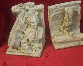 May include: Two off-white bookends with a musical theme. One features a bust of a man with a powdered wig, the other a scroll with musical notes. Both are decorated with floral accents.