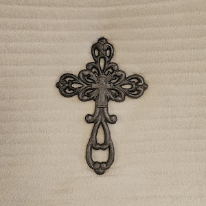 Könnte beinhalten: Verziertes, dunkelgraues Metallkreuz mit filigranen Details. Das Kreuz hat ein dekoratives Design mit einer Figur in der Mitte und einem ausgestellten Sockel. Es liegt auf einem strukturierten, hellbeigen Hintergrund.
