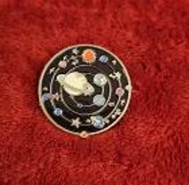 May include: A round, black enamel pin featuring a gold-colored planet and orbiting planets with colorful accents. The pin has a gold-colored border and is set against a red, textured background.